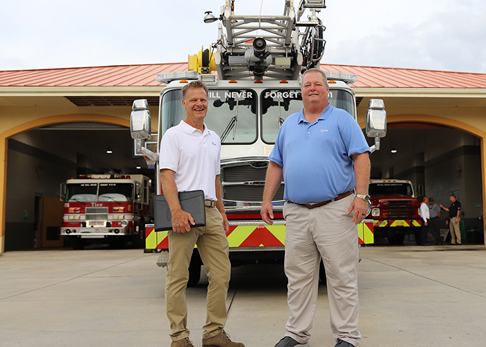 photo of VFIS associate and client standing in front of a fire engine that is parked in front of a fire hall and has open bays with parked firetrucks in the background