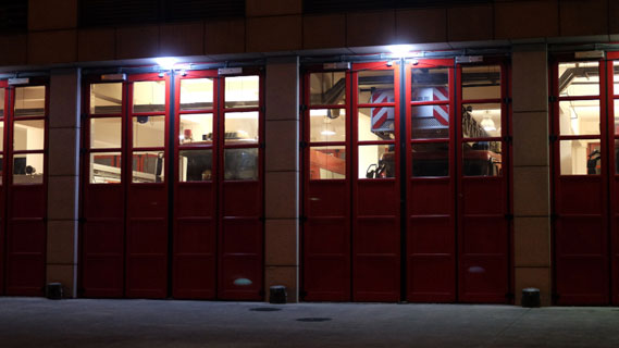 The front of a firestation with all of the bays closed at night, firetrucks visible through the windows